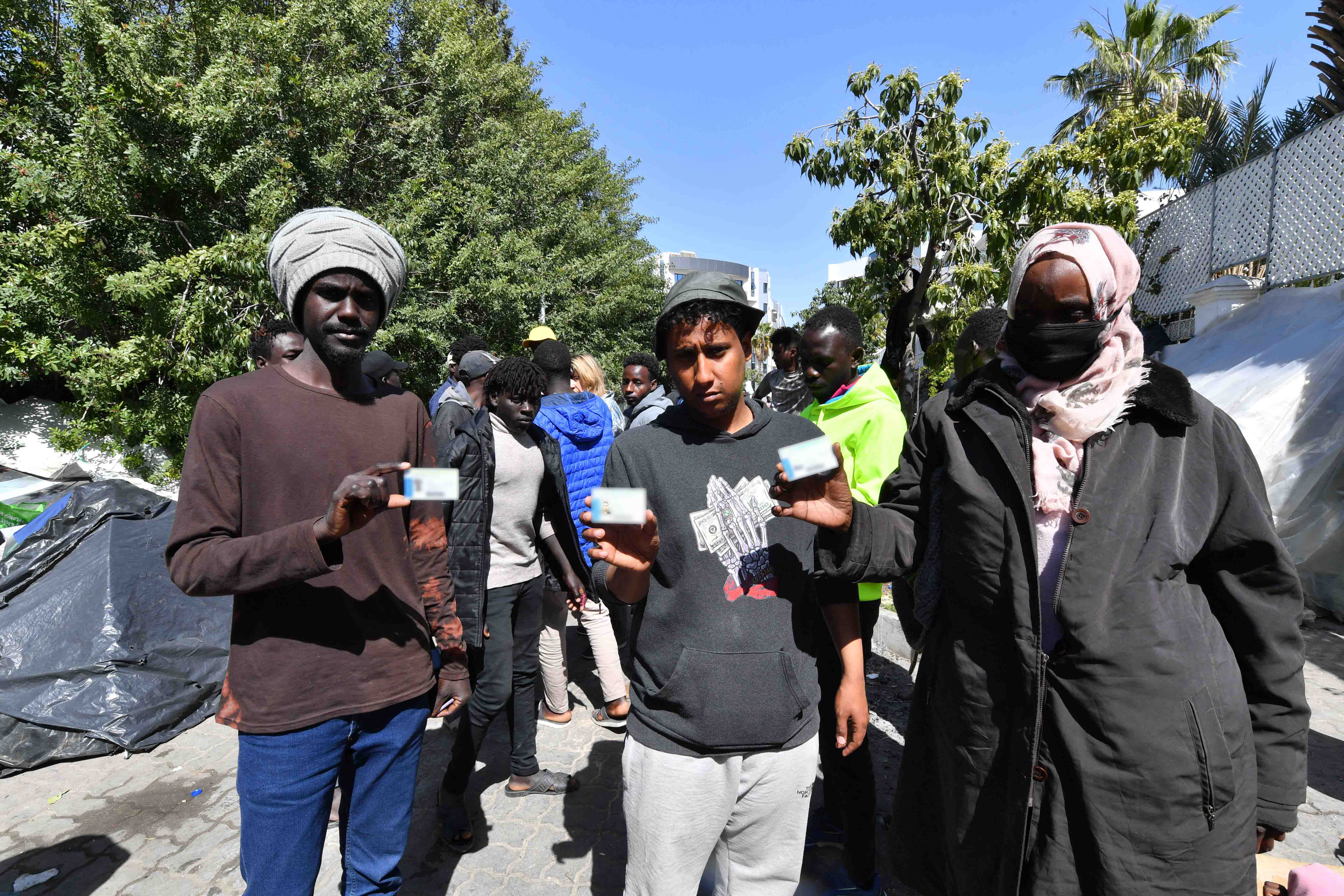 Des hommes originaires du Soudan et du Yémen montrent leur carte de réfugié ou de demandeur d’asile le 12 avril 2023 devant le bureau de l’Organisation internationale pour les migrations (OIM) à Tunis. Le 11 avril, la police tunisienne avait fait usage de la force et de gaz lacrymogènes pour disperser des réfugiés, des demandeurs d’asile et des migrants siégeant devant le bureau de l’Agence des Nations Unies pour les réfugiés (UNHCR) à Tunis dans un camp de fortune qui a ensuite été démantelé. © 2023 Fethi 