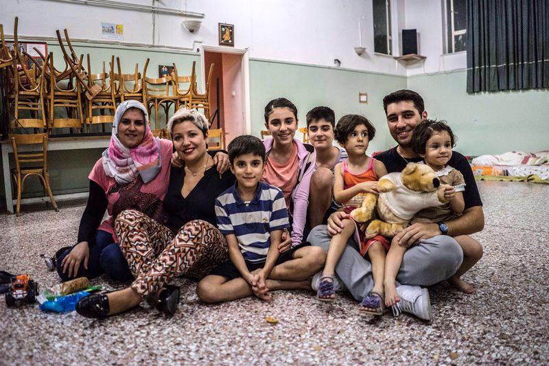 Falak from Syria – with her family Elin (2), Evin (4), Rudi (10), Mohamed (11) and Lava (13) – and Sperantza from Greece with her son Alexandros pose for a family photo in the temporary accommodation provided by the local Catholic parish in Aigaleo, Athen