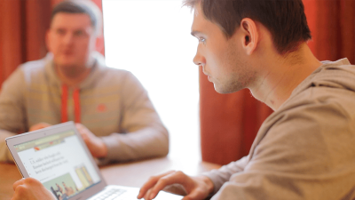 A young man in a gray hoodie, Ruslan Sokolovsky, sits with his laptop, with his lawyer in the background